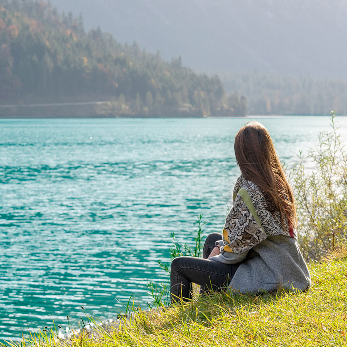 Person sitzt am Ufer eines klaren Sees und blickt auf das Wasser 
