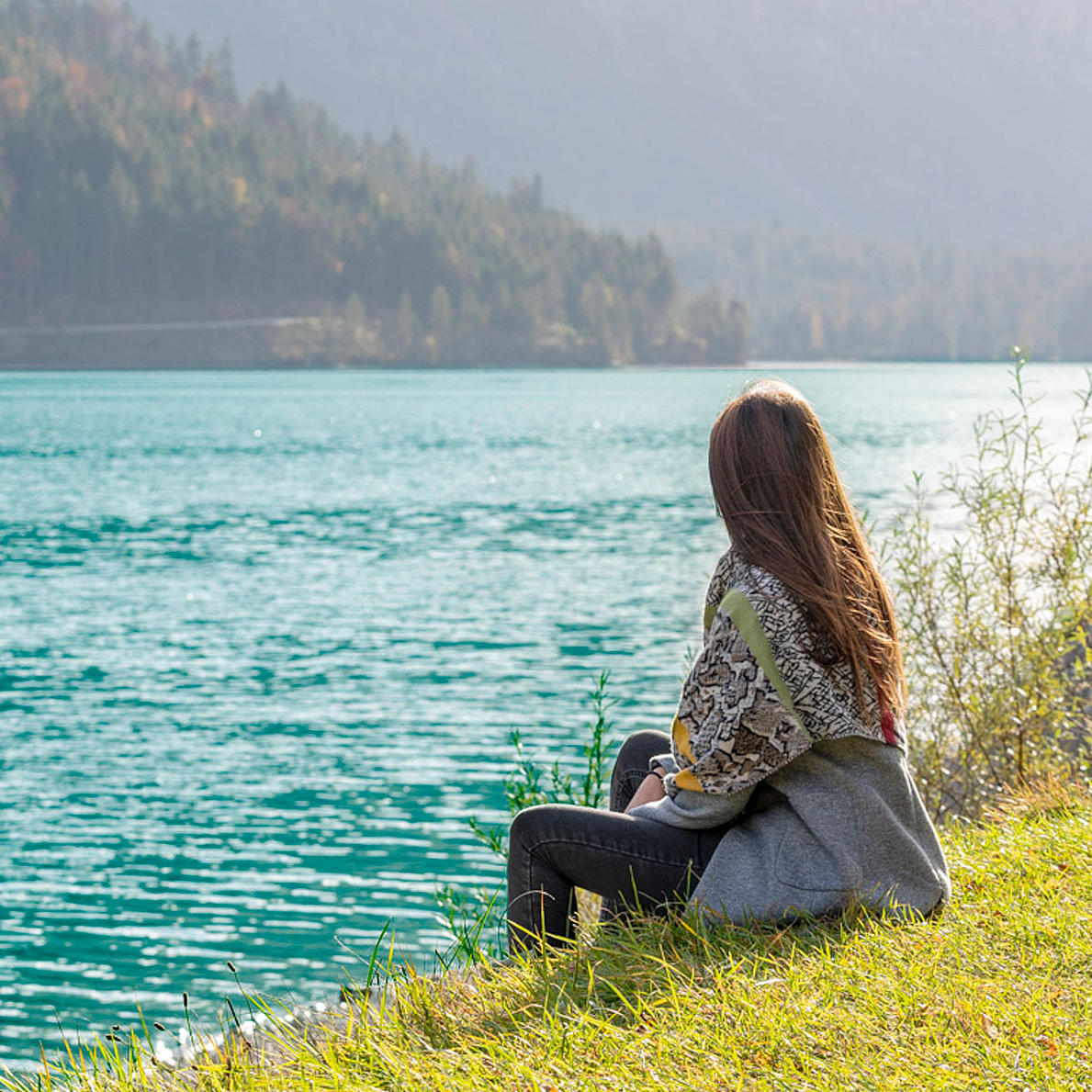 Person sitzt am Ufer eines klaren Sees und blickt auf das Wasser 