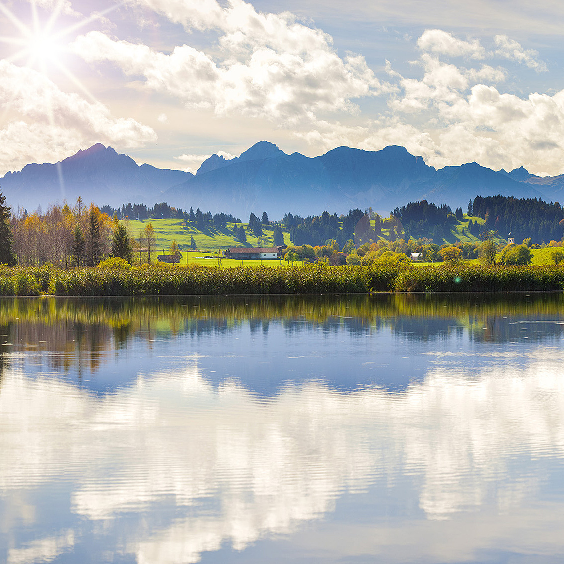Spiegelung von Landschaft, Bergen und Sonne in ruhigem See im Allgäu