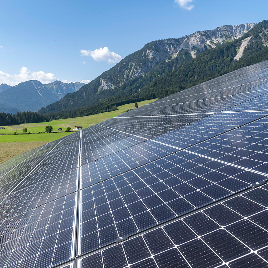 Photovoltaik-Anlage auf Dach im Vordergrund, im Hintergrund Blick auf Felder und Bergkulisse