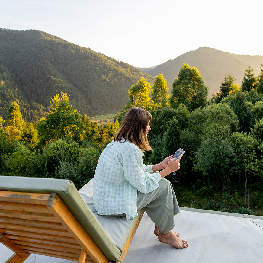 Frau sitzt mit Handy auf einem Liegestuhl im Freien mit Ausblick auf einen Wald und Berge